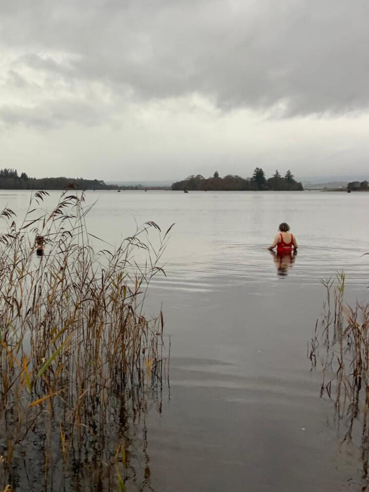 A photo of Lisa, a white woman, wearing a red swimsuit , waist high in a lake in autumn. There are pine trees in the distance.