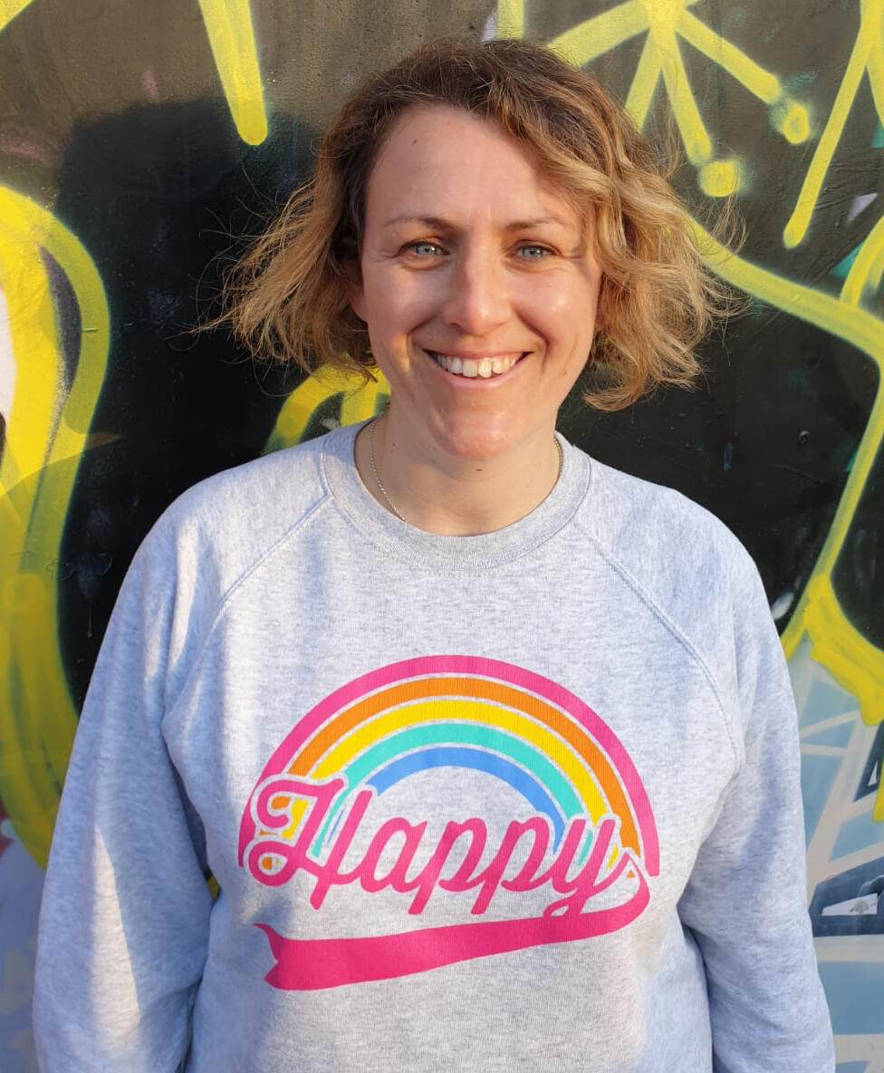 White woman in a sweatshirt that says 'happy' is standing and smiling against the backdrop of a wall with black and yellow graffiti on it